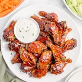 Chicken wings on a white serving platter with a small bowl of creamy dip. Platter is flanked by bowls of baby carrots and celery pieces.