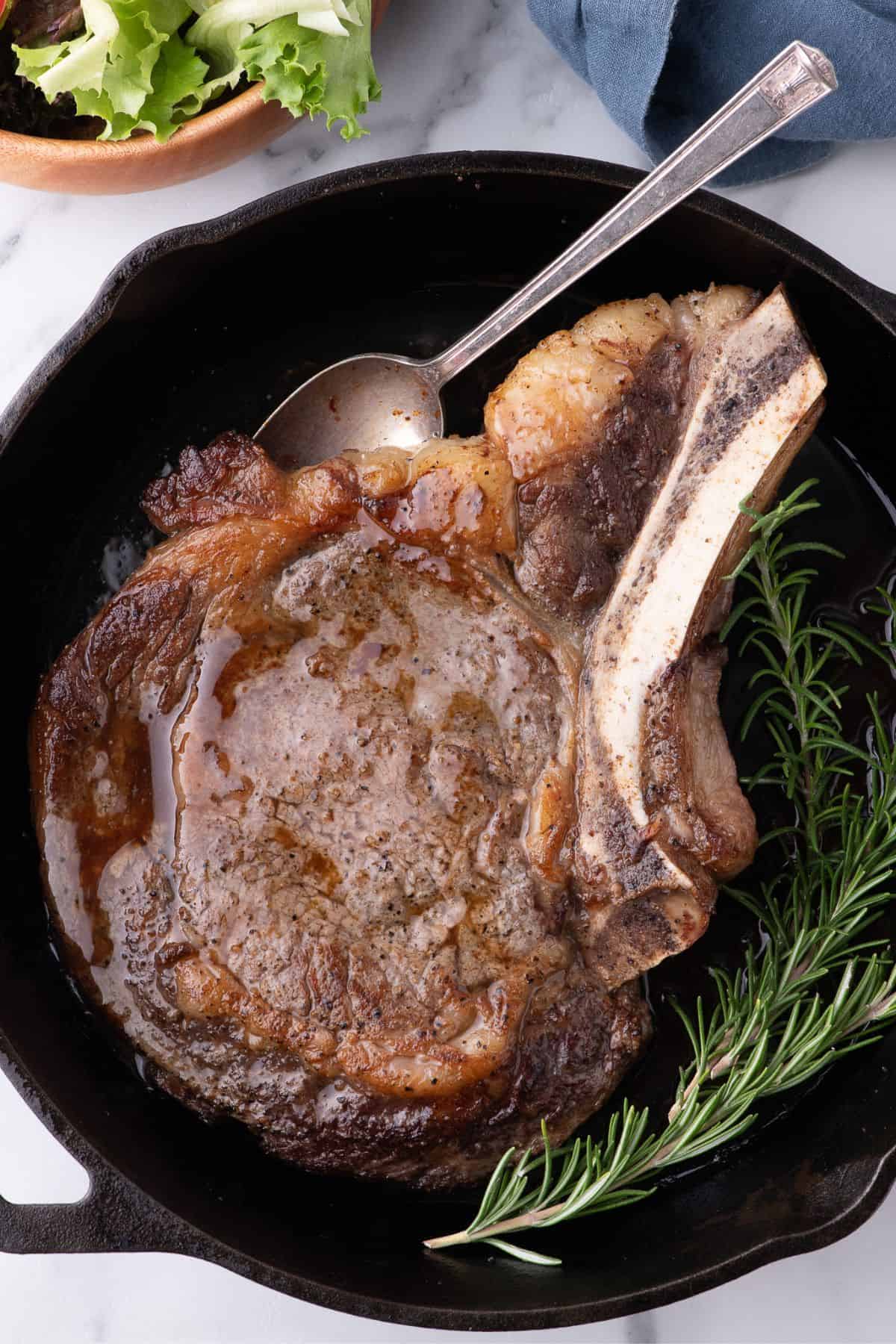 Completed ribeye steak in a cast iron pan with a sprig of rosemary.