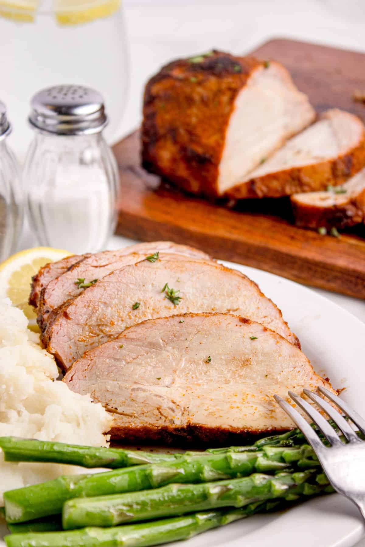 Grilled pork loin on a white dinner plate with asparagus and mashed potatoes. A wooden cutting board is behind it wit the rest of the sliced pork loin.