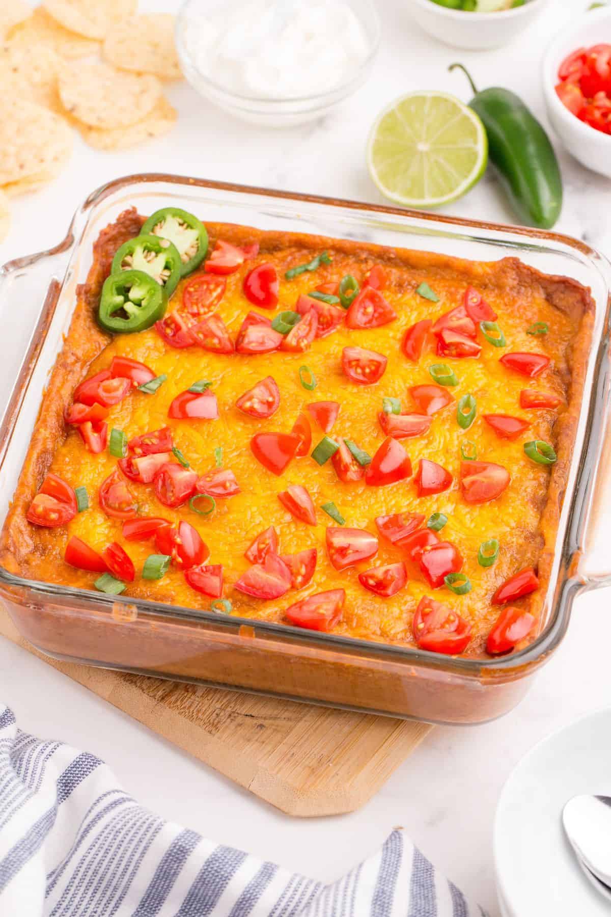 Completed refried bean dip in a glass baking pan. Dip has been garnished with diced tomatoes, green onions, and sliced jalapenos.