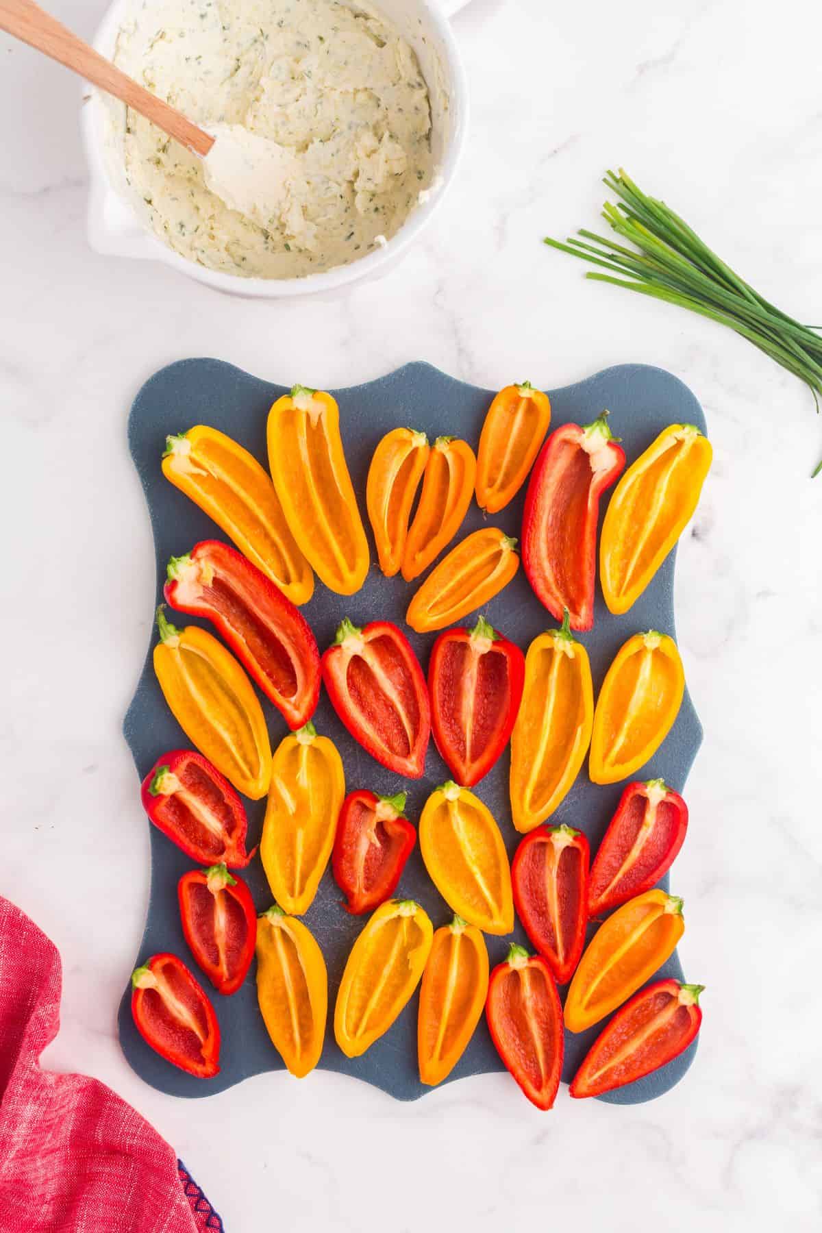 Peppers are halved on a medium sized cutting board. A mixing bowl with the cream cheese mixture is sitting above the cutting board with a spatula in it.