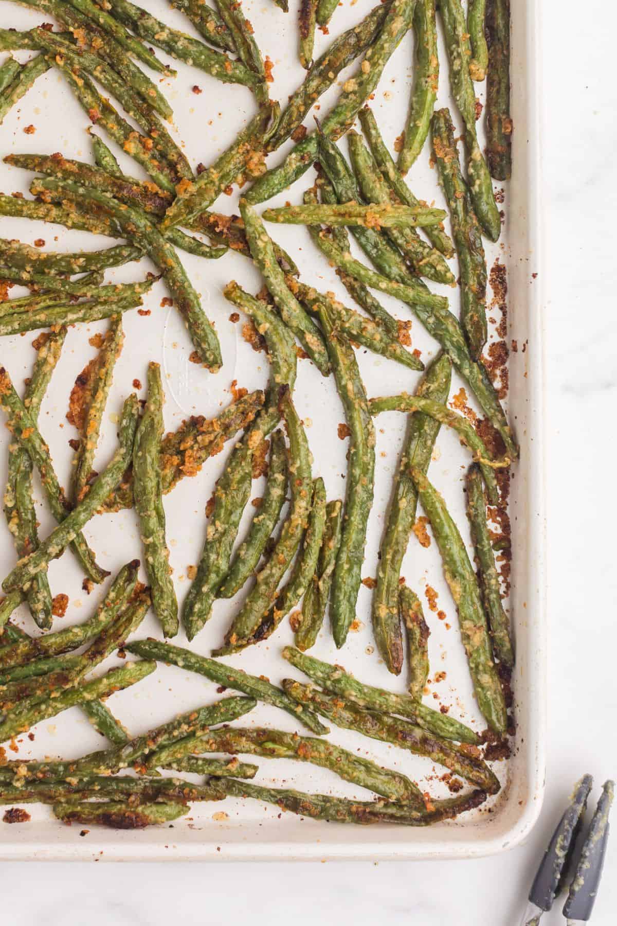 Baked green beans on a baking sheet. A pair of tongs sit to the side of the sheet pan.