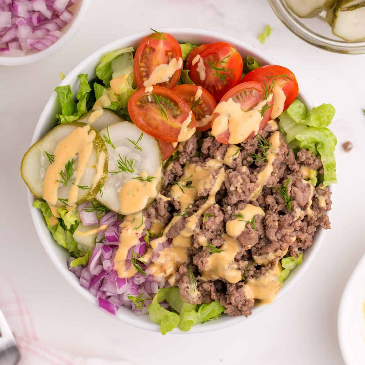 Burger bowl from an overhead view with ground beef, sauce, lettuce, tomatoes, pickles, and more.