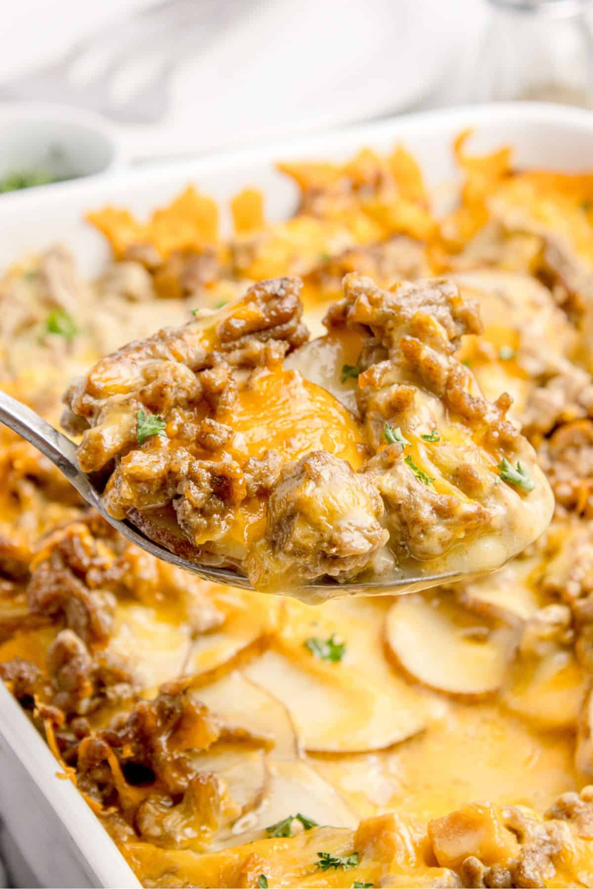 A serving spoon lifting up a portion of cheesy hamburger potato casserole above the baking dish.