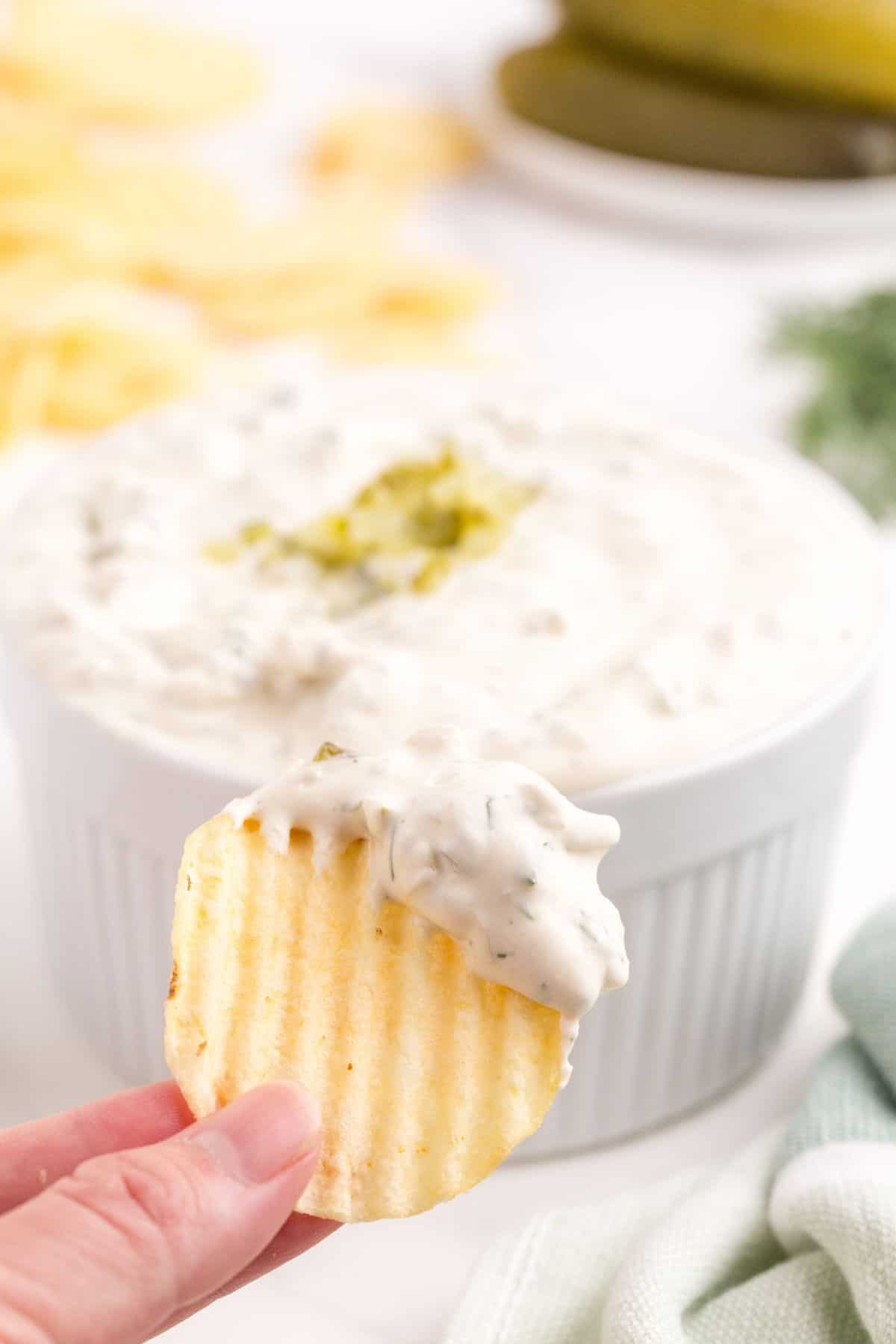 A hand is holding a dipped ridged potato chip in front of a ramekin with the completed dill pickle dip.