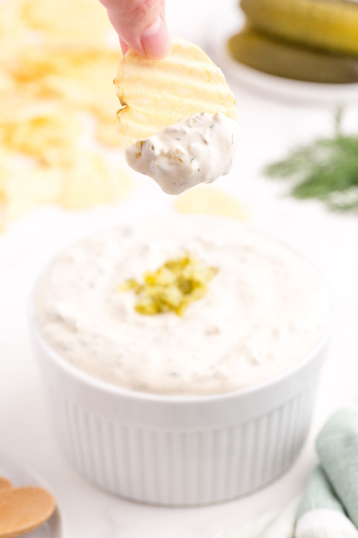 Completed dill pickle dip in a ramekin. A hand has dipped a redged potato chip in the dip and is positioned above the cup.