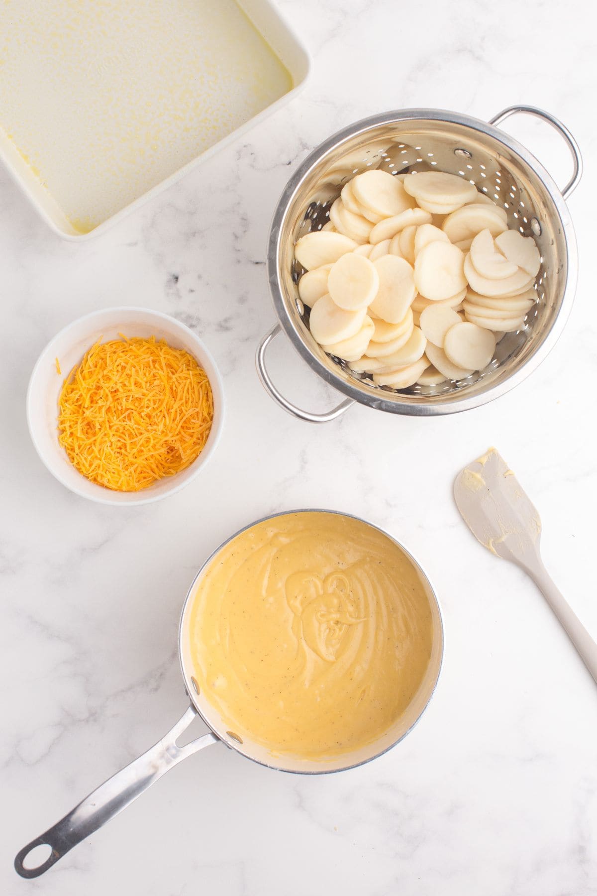 Completed cheese sauce in a small saucepan. Next to the saucepan is a colander holding the drained canned potatoes, and a white bowl with the remaining shredded cheese.