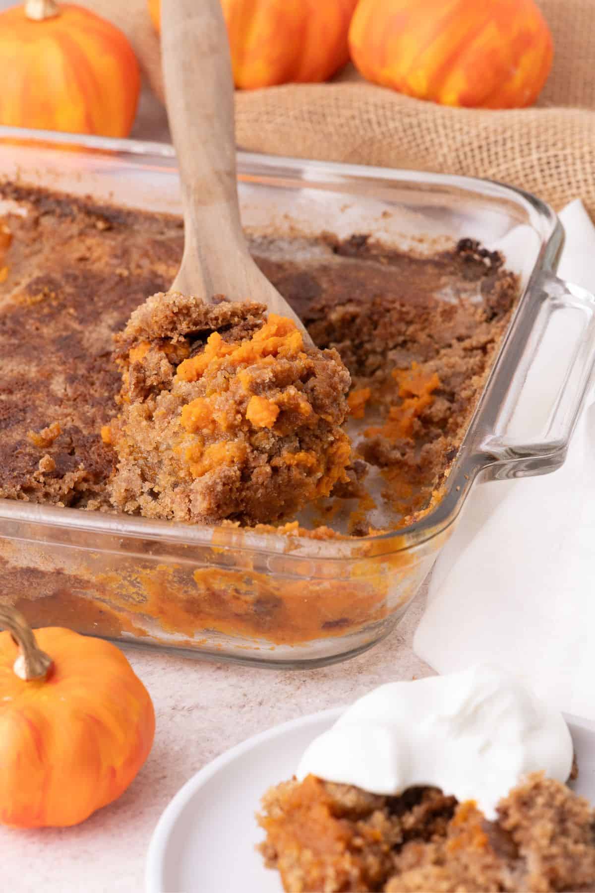 Completed pumpkin dump cake in a large, clear baking pan. A wooden spoon is scooping a serving out the pan.