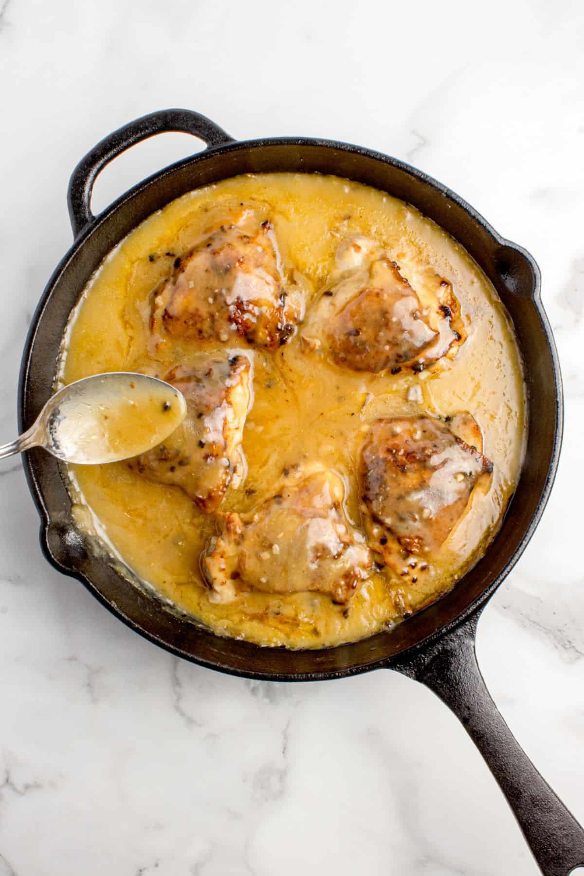 Chicken thighs in a cast iron pan with gravy. A spoon is pouring the gravy over the tops of the chicken.