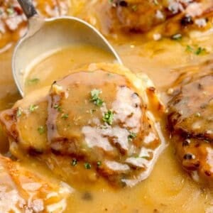 Chicken thighs in a cast iron pan with gravy. A large serving spoon is scooping up a thigh.