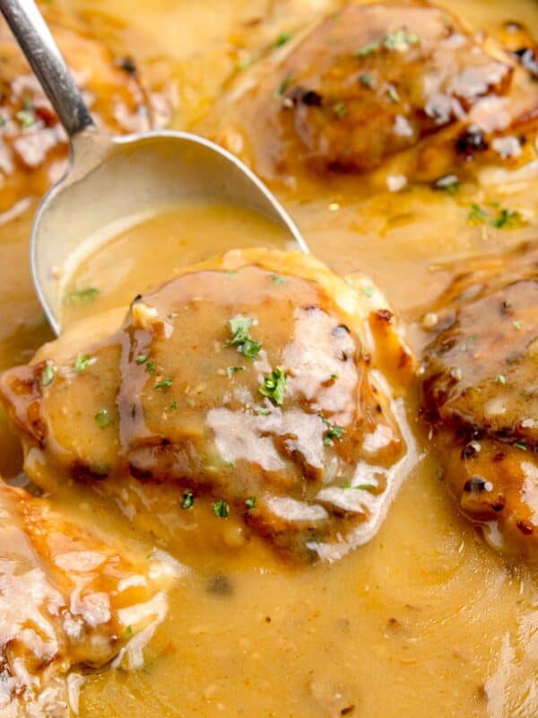 Chicken thighs in a cast iron pan with gravy. A large serving spoon is scooping up a thigh.