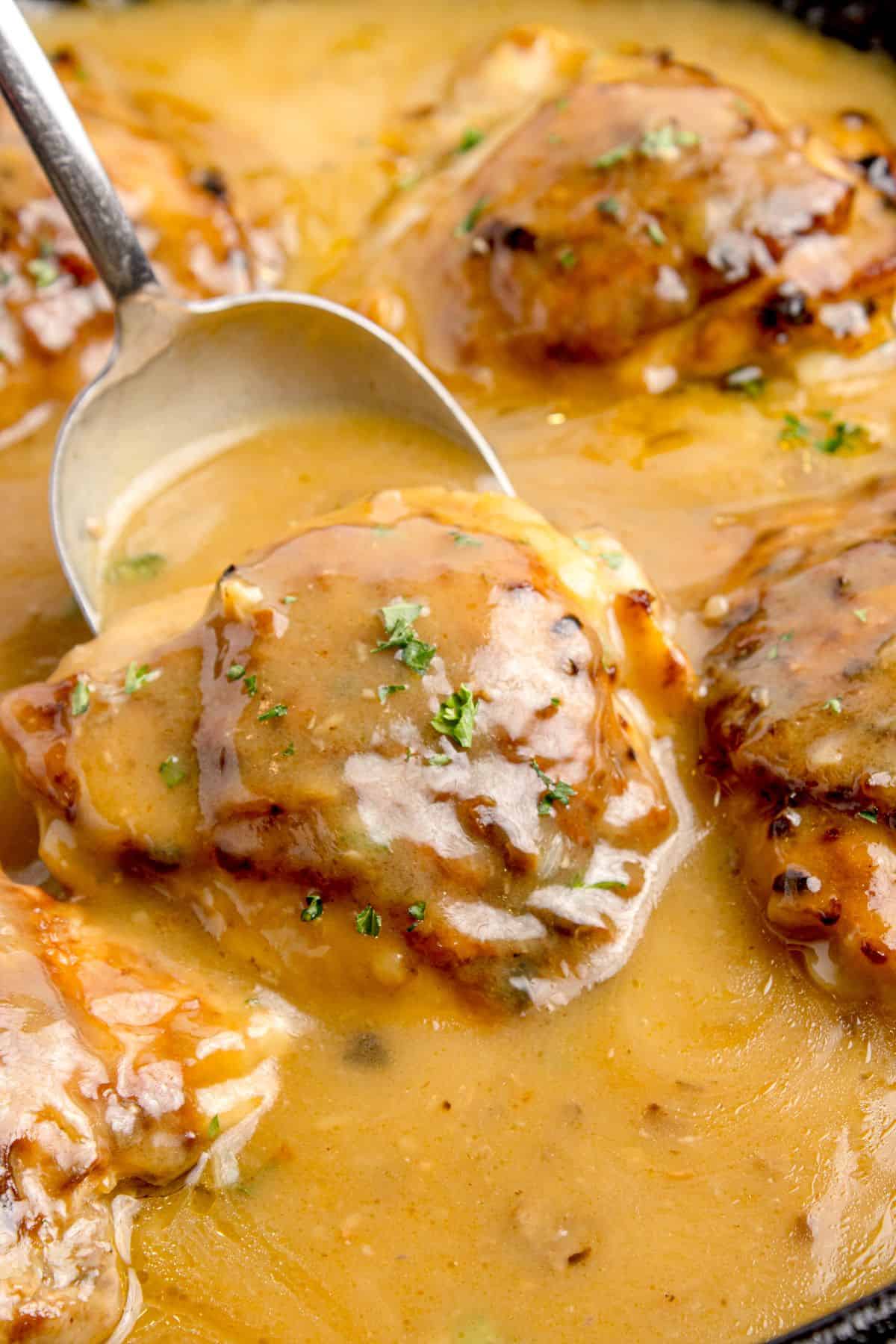 Chicken thighs in a cast iron pan with gravy. A large serving spoon is scooping up a thigh.