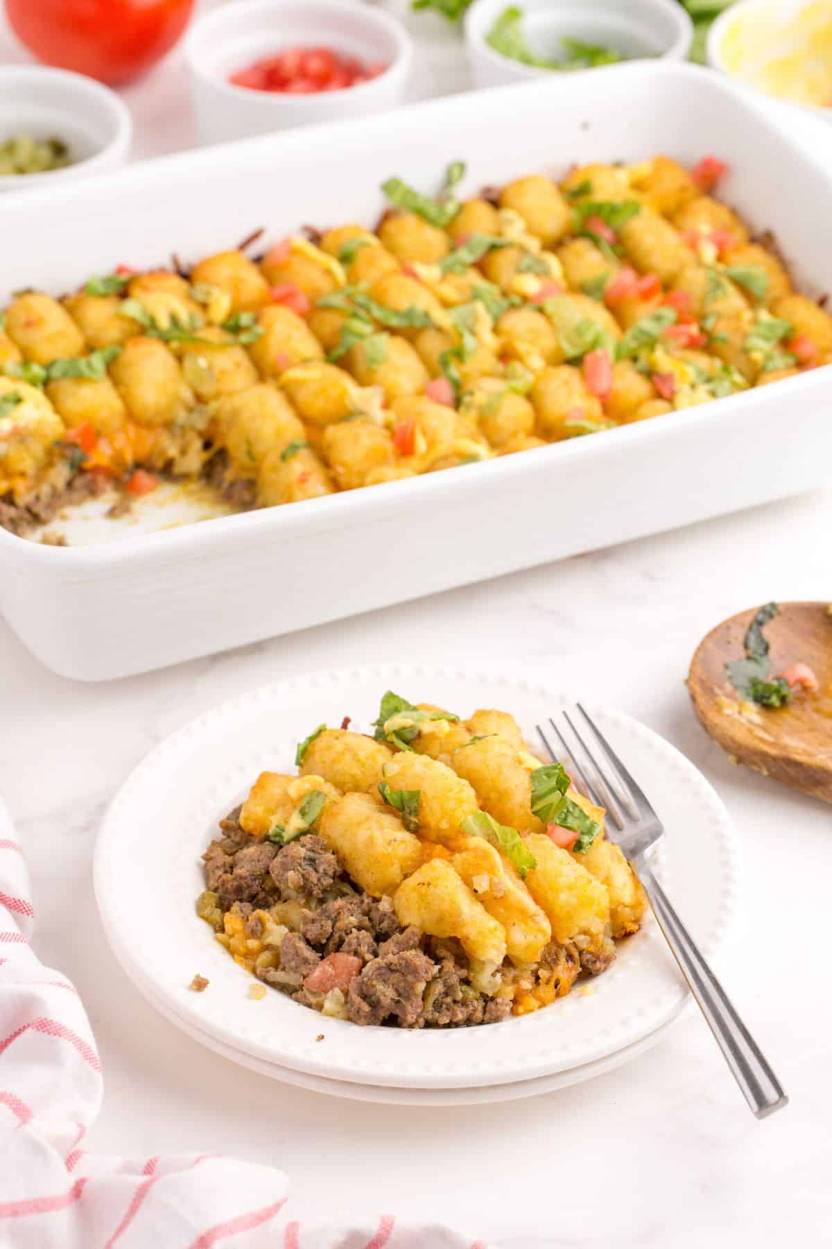 Big mac casserole on a white dinner plate. A white baking dish with the rest of the casserole is placed behind the dinner plate.