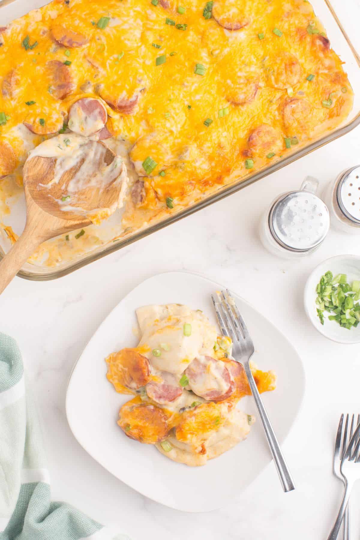 A serving of pierogi casserole on a white dinner plate. The remaining of the casserole is in a glass baking pan next to the dinner plate.