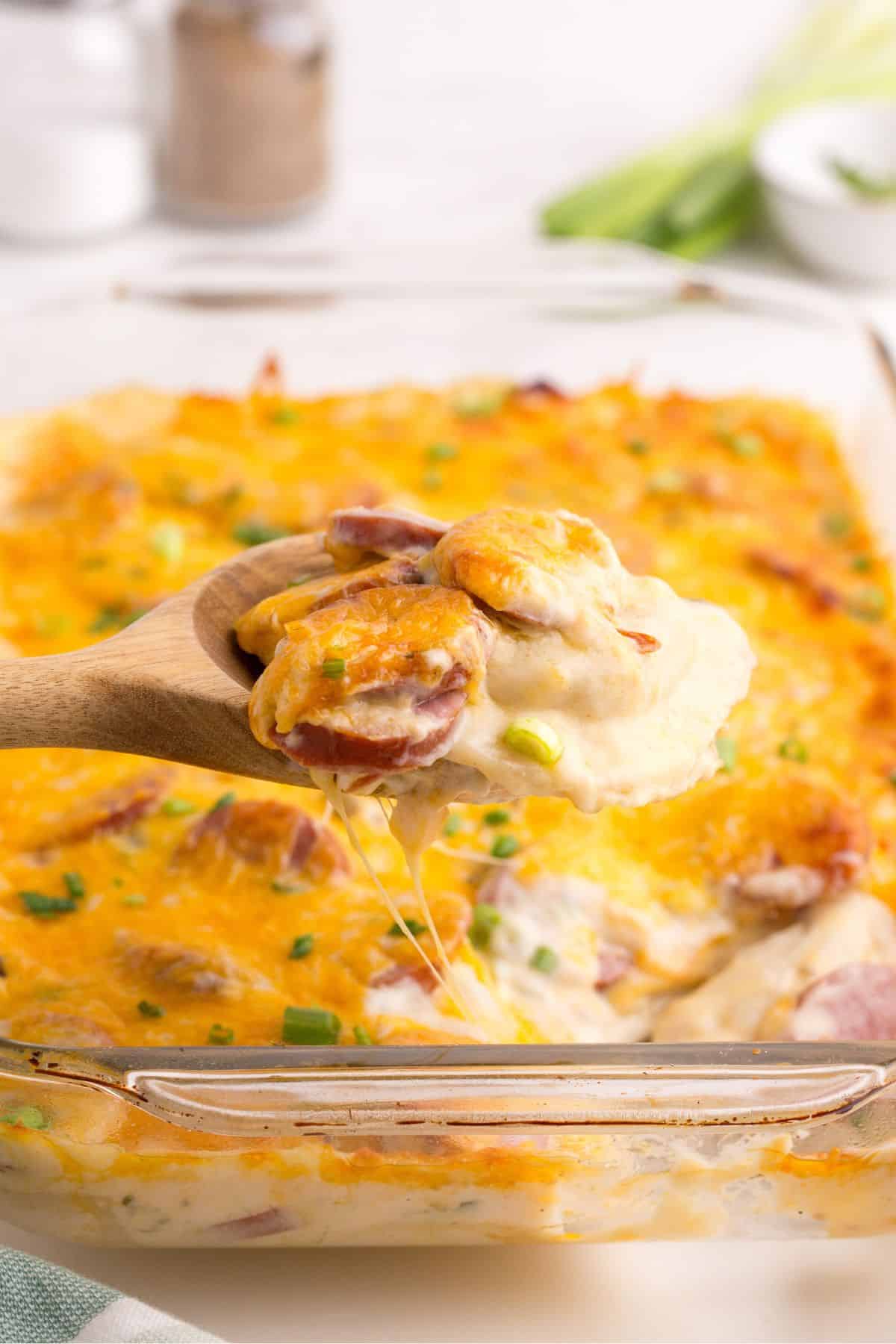 Pierogi casserole in a glass baking dish. A wooden serving spoon is lifting a serving out of the pan.