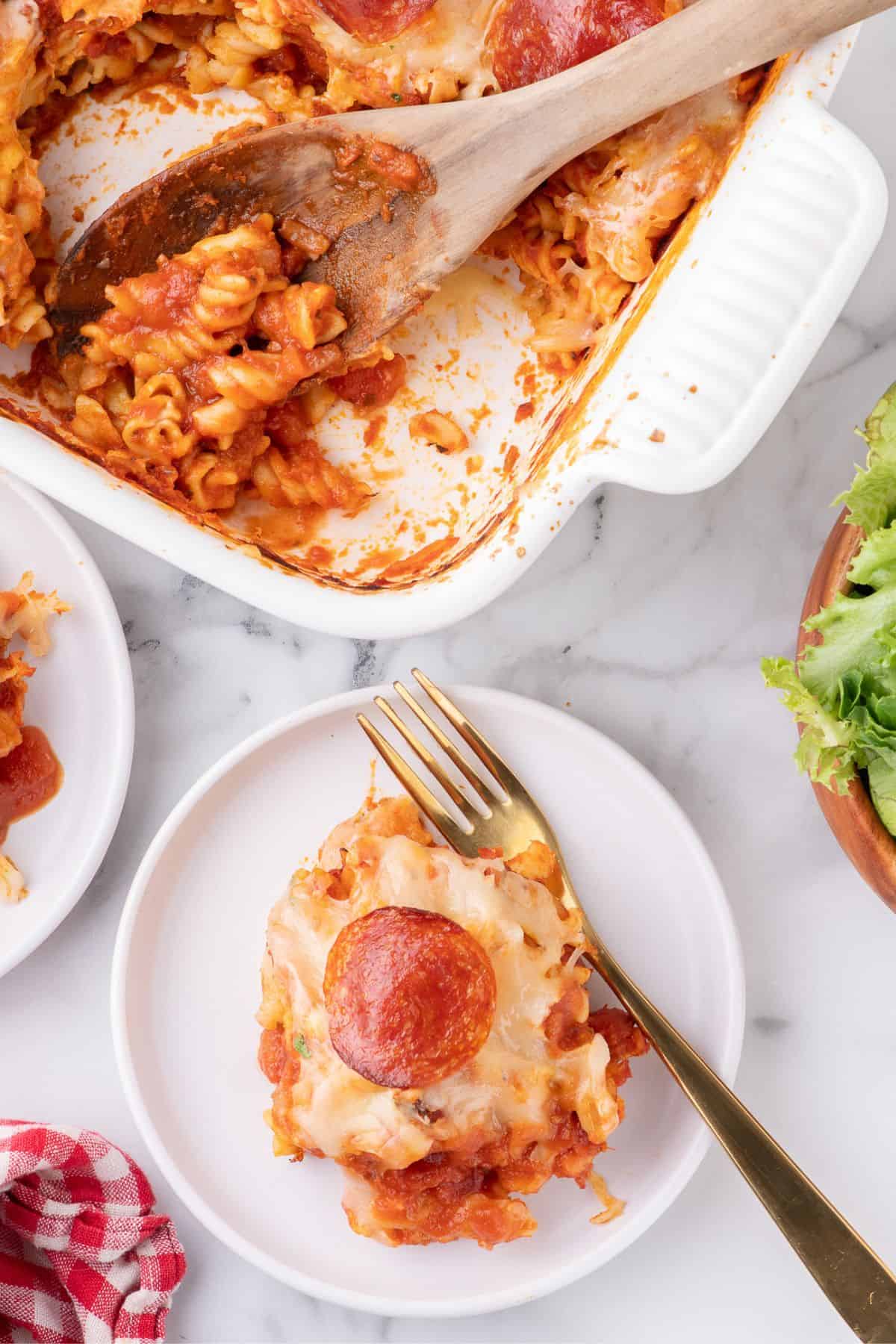 A white dinner plate with a serving of pizza casserole on it. The remainder of the casserole is in a white baking dish just behind the dinner plate.