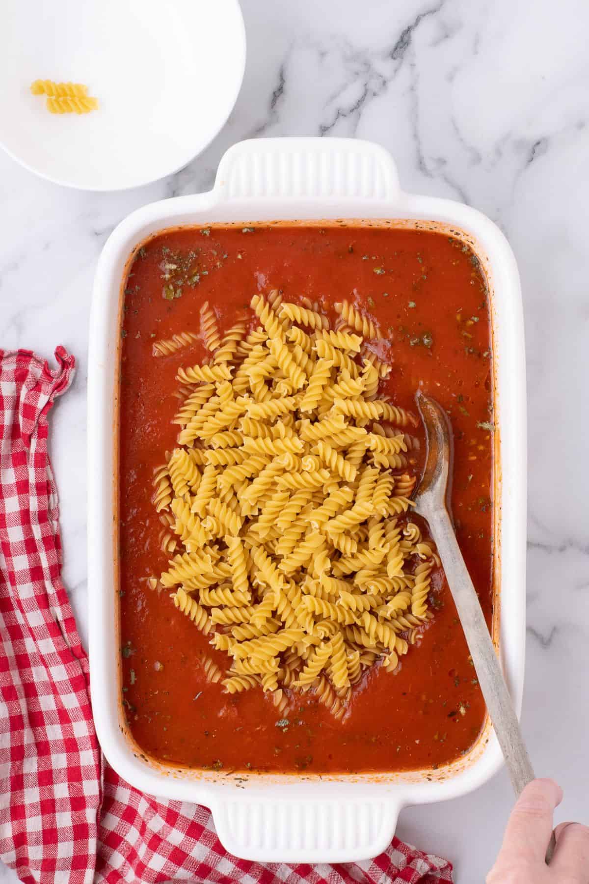 Pasta added to the sauce in the white baking dish.