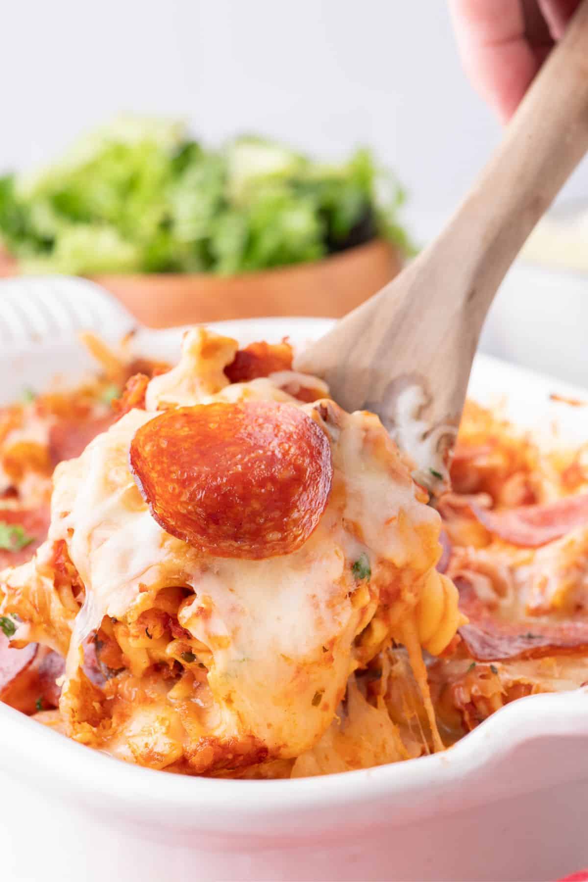 A wooden spoon scooping up a serving of pizza casserole from a white baking dish.