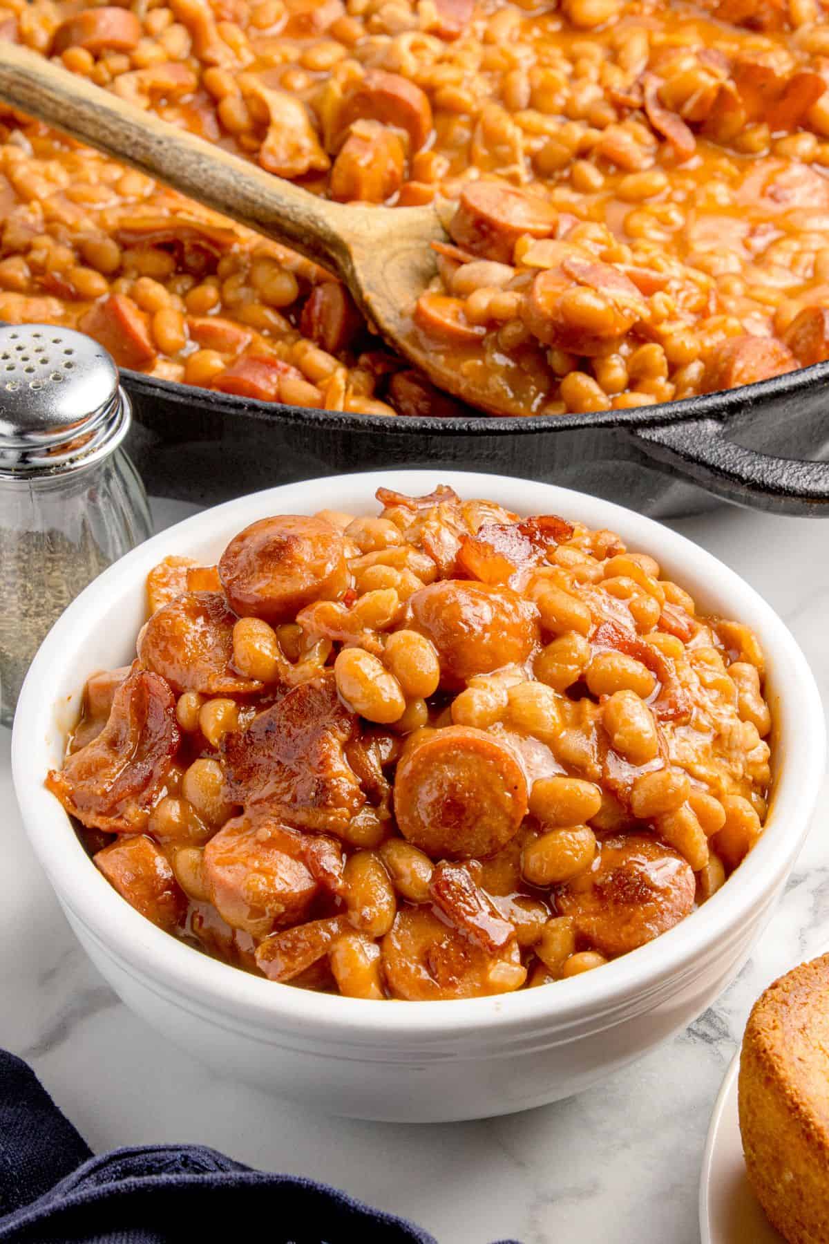 Completed beanie weenies in a white bowl. A cast iron pan with the remainder of the recipe sits behind the bowl.