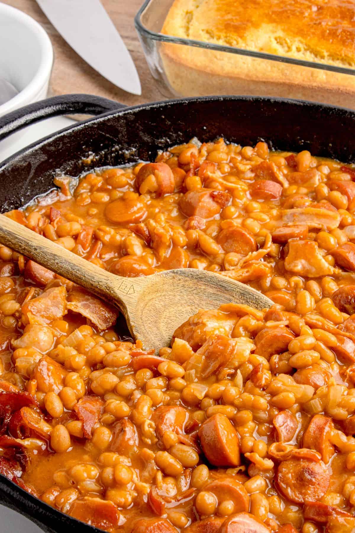 Beanie weenies recipe in a cat iron pan with a wooden spoon.