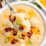 Completed cheesy potato soup in a white bowl, garnished with shredded cheese, green onion, and bacon bits. A spoon is lifting a bite above the bowl.
