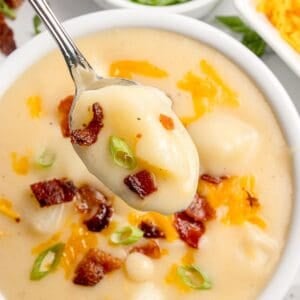 Completed cheesy potato soup in a white bowl, garnished with shredded cheese, green onion, and bacon bits. A spoon is lifting a bite above the bowl.