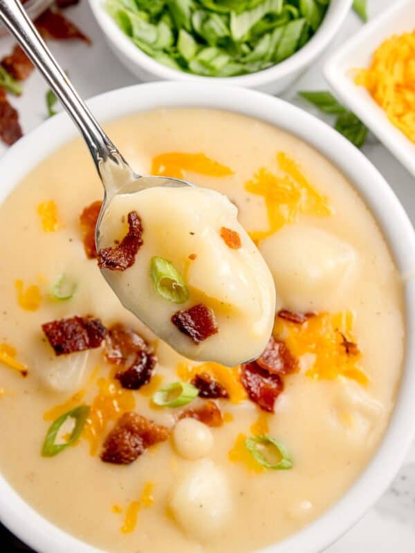 Completed cheesy potato soup in a white bowl, garnished with shredded cheese, green onion, and bacon bits. A spoon is lifting a bite above the bowl.