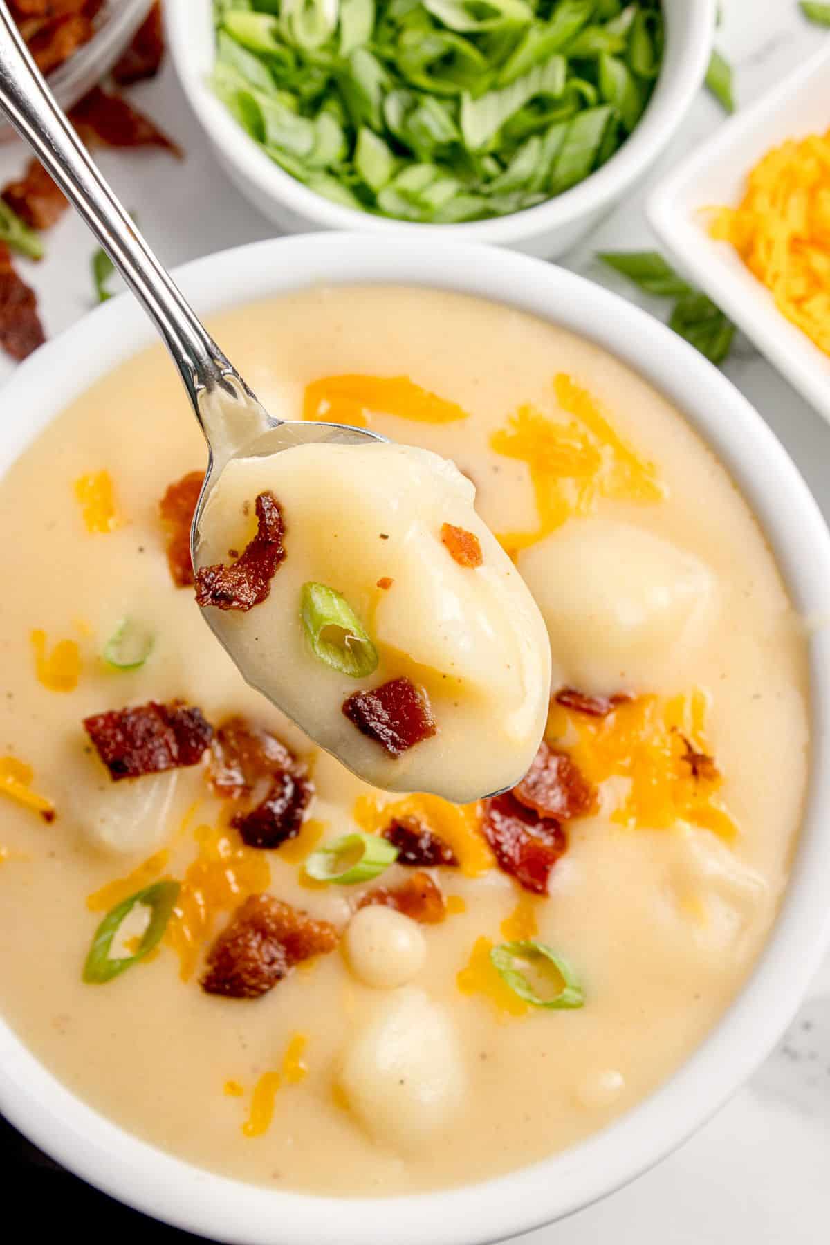 Completed cheesy potato soup in a white bowl, garnished with shredded cheese, green onion, and bacon bits. A spoon is lifting a bite above the bowl.