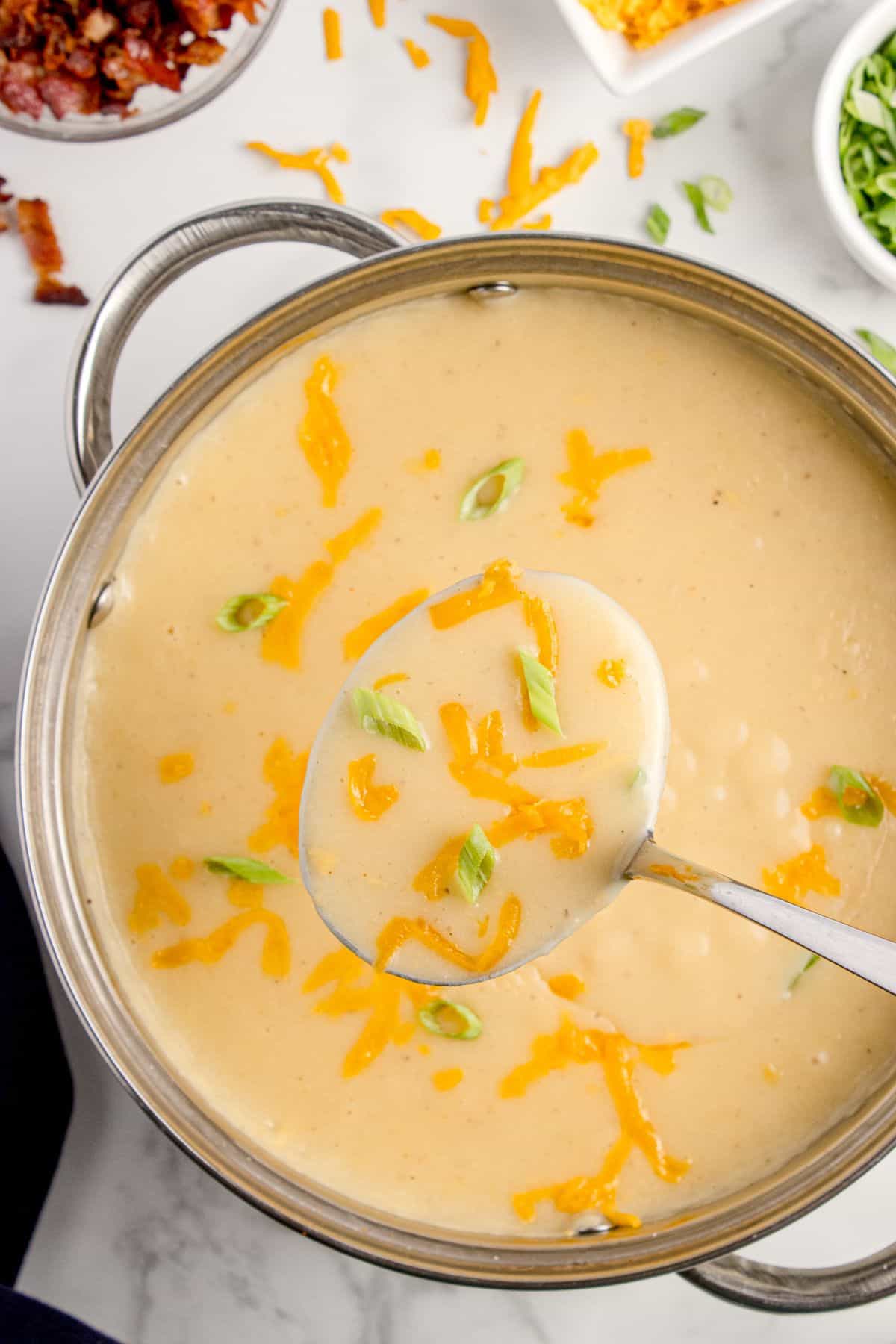 A pot of cheesy potato soup garnished with green onions and shredded cheddar cheese. A ladle is lifting a serving out of the pot.