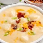 Potato soup in a white bowl. It is garnished with green onions, cheddar cheese, and bacon.