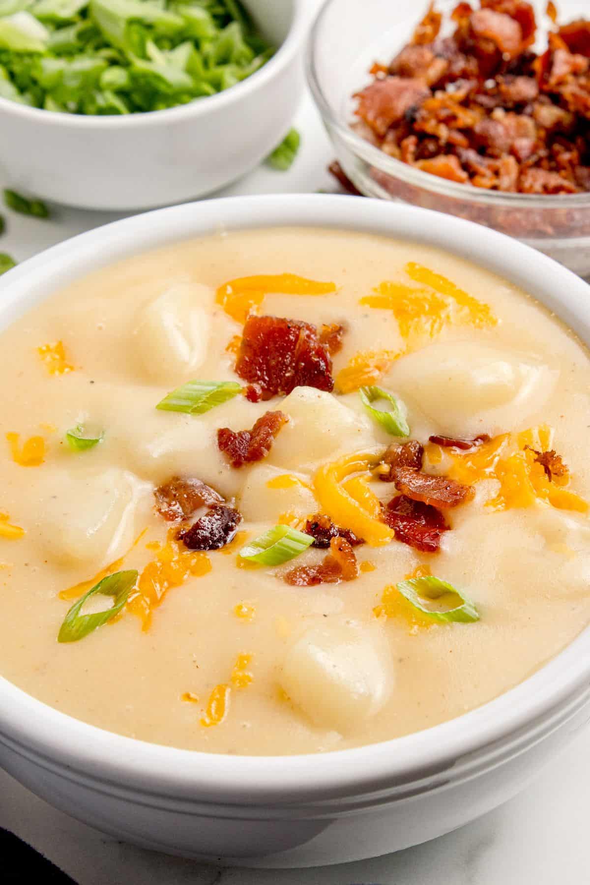 Potato soup in a white bowl. It is garnished with green onions, cheddar cheese, and bacon.