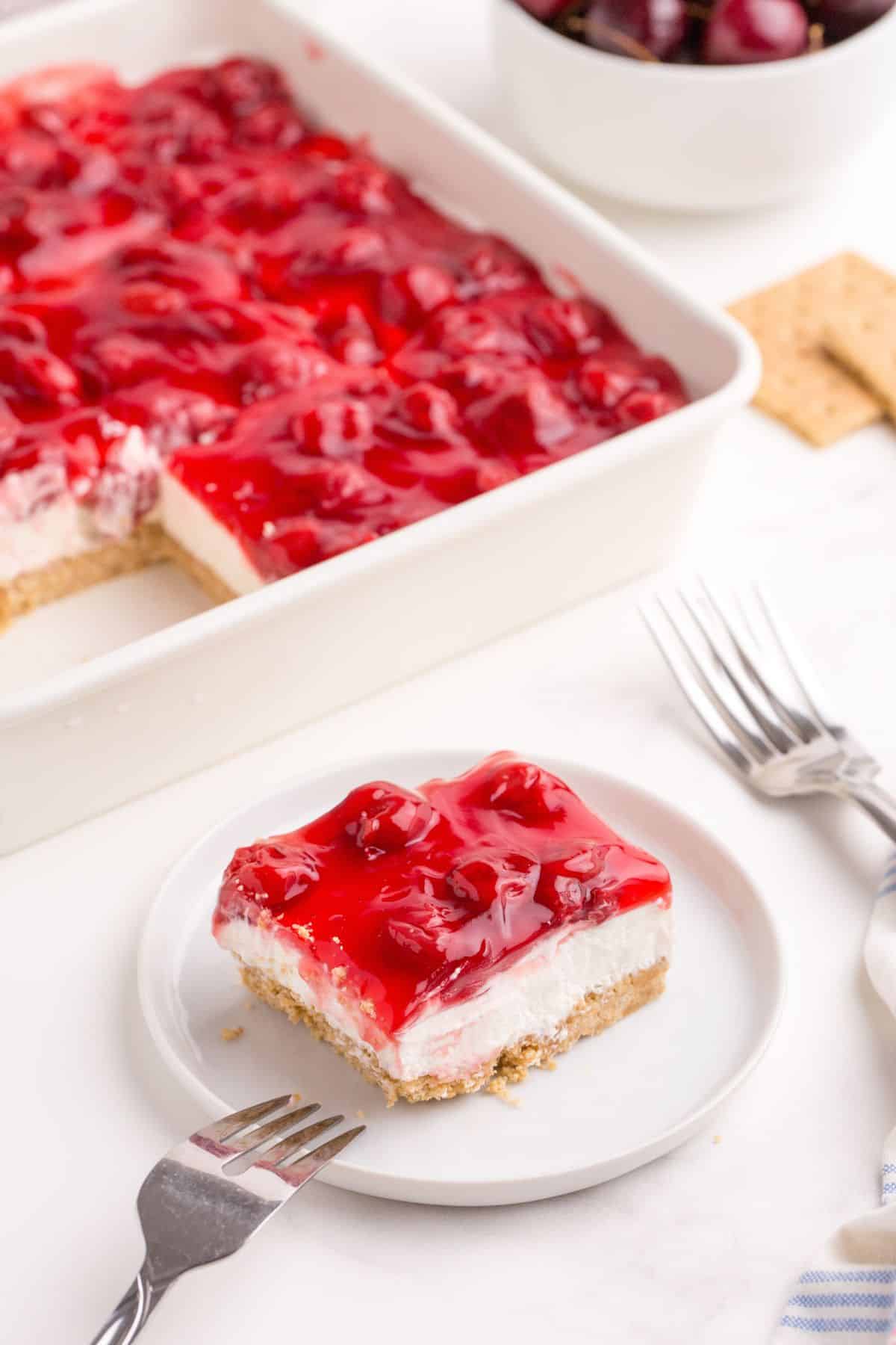 Completed cherry delight dessert. A white plate has a slice of cherry delight on it. Behind the plate is a square baking pan with the remainder of the dessert in it.