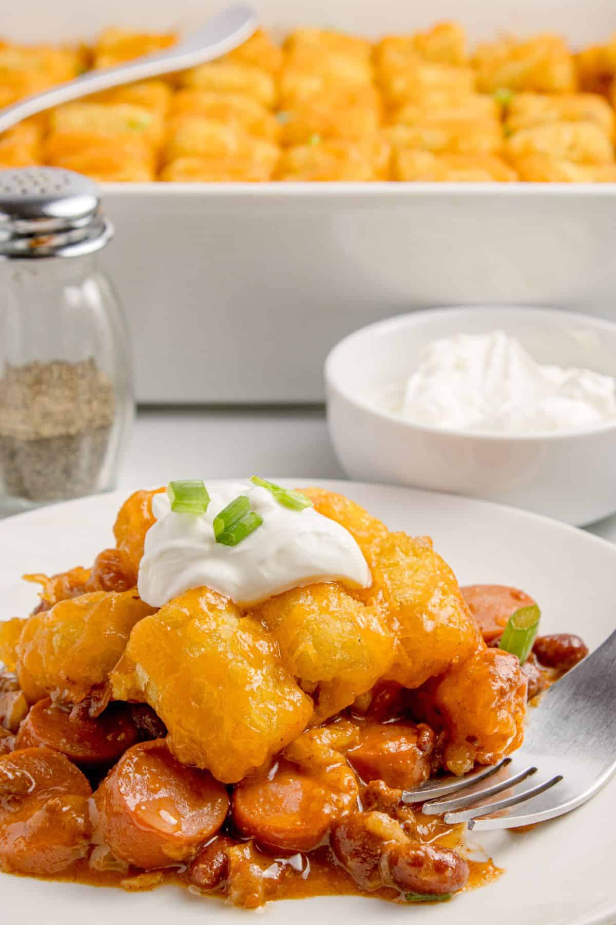 A serving of hot dog casserole on a white dinner plate. Behind the plate is a white baking dish with the remaining hot dog casserole.
