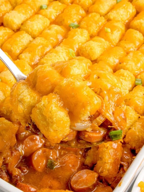 A serving spoon lifting a portion of hot dog casserole out of the baking dish.