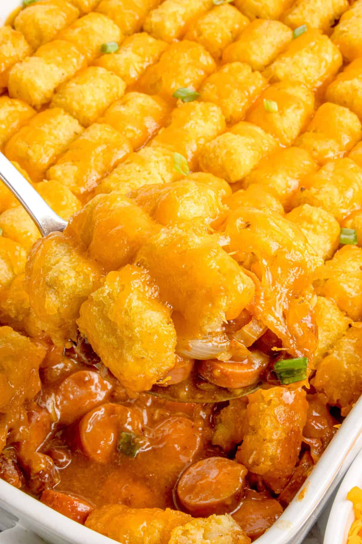 A serving spoon lifting a portion of hot dog casserole out of the baking dish.