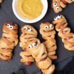 Six hot dog mummies placed around a small bowl of mustard.