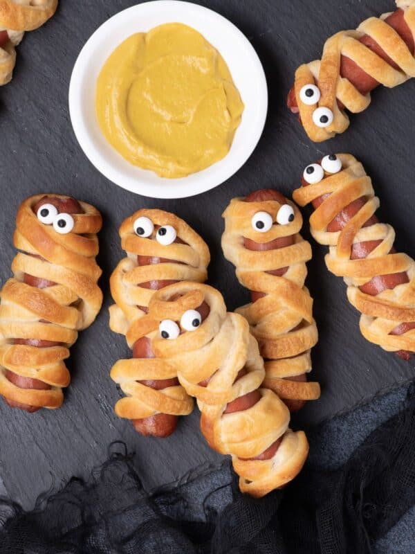 Six hot dog mummies placed around a small bowl of mustard.