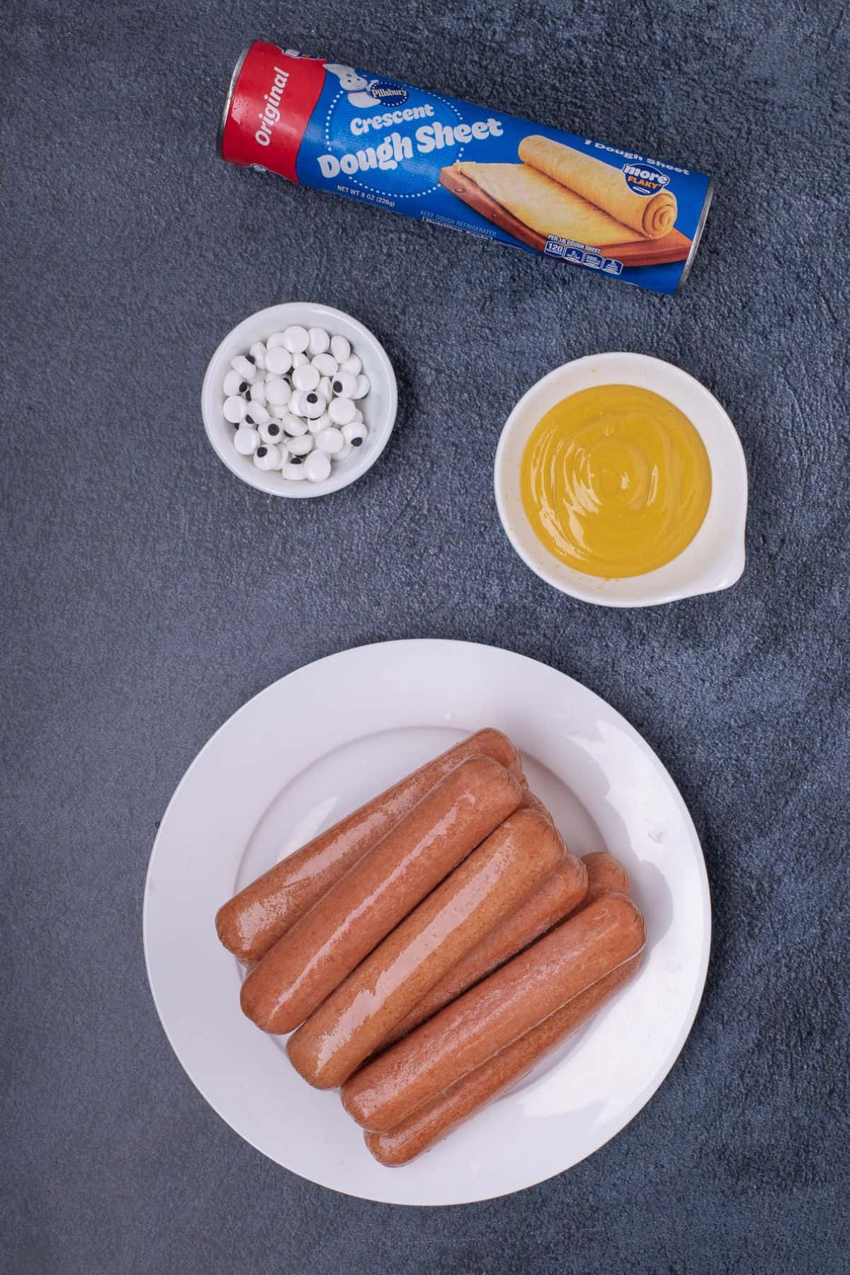 Ingredients for hot dog mummies: crescent roll dough, mustard, hot dogs, and candy eyes.