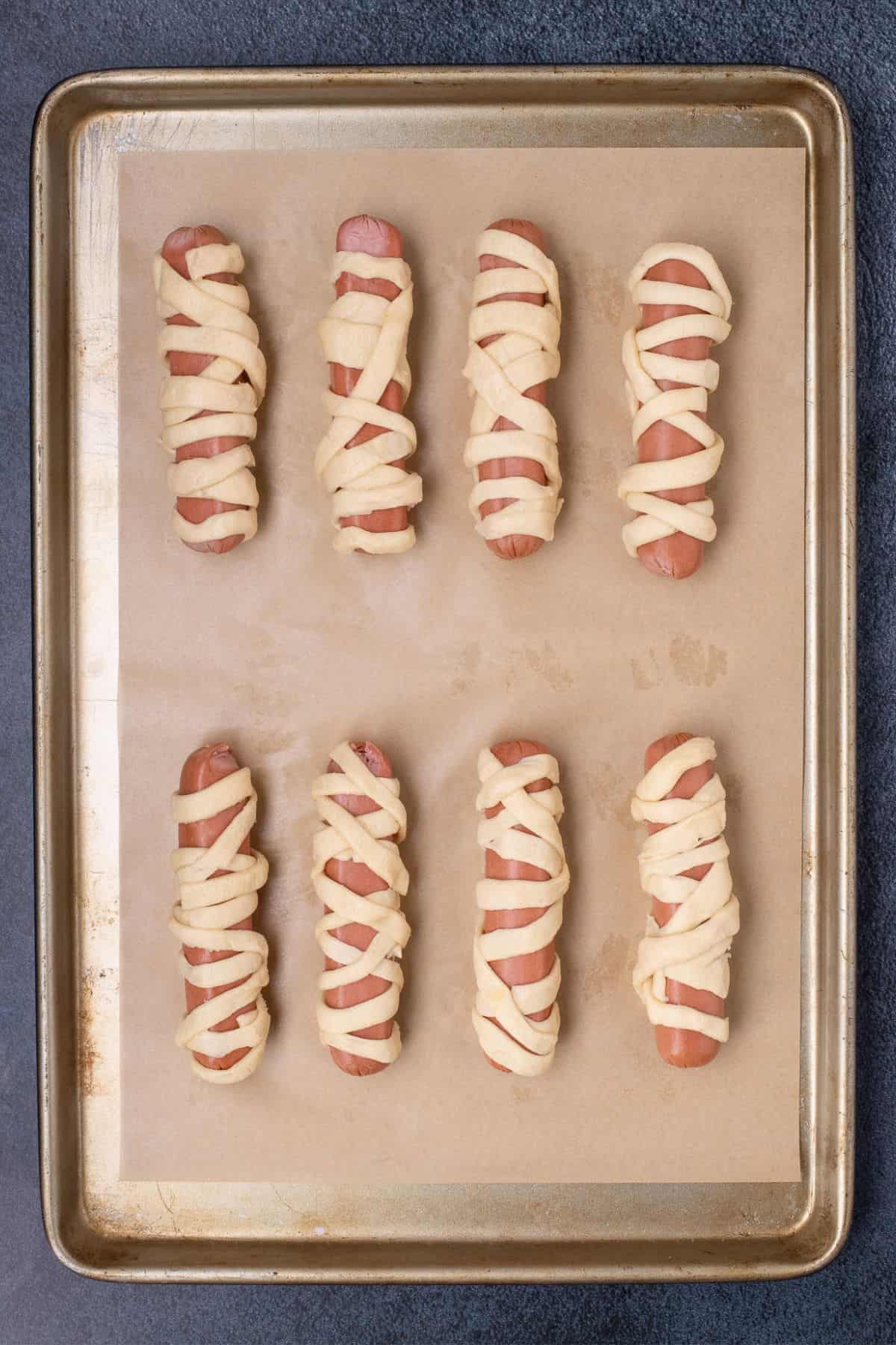 Raw crescent dough strips wrapped around hot dogs on a parchment lined baking sheet.