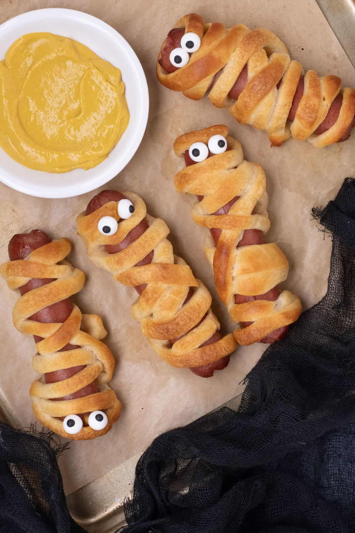 Four hot dog mummies arranged around a small bowl of mustard.