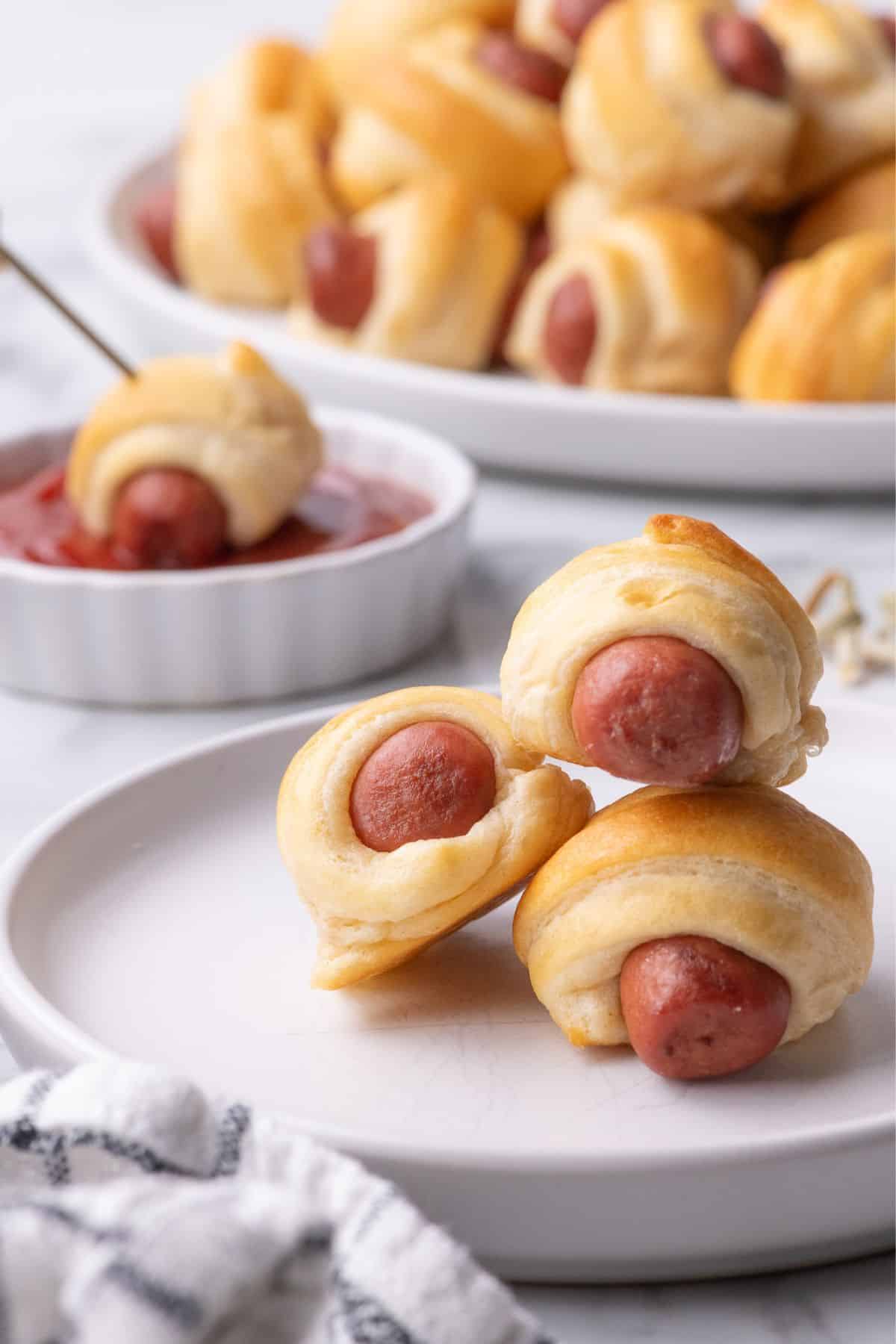 Pigs in a blanket stacked up on a white plate. Behind the plate a single pig in a blanket is in a ramekin of ketchup.