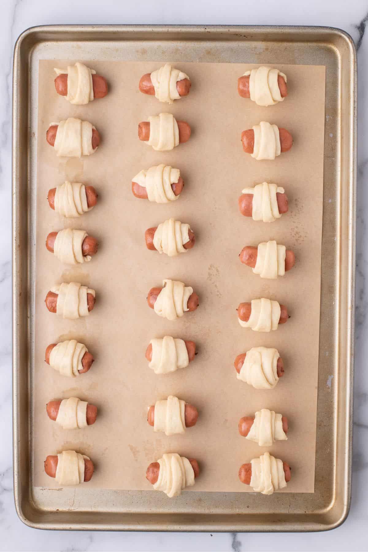 Pigs in a blanket are arranged on a parchment lined baking sheet.