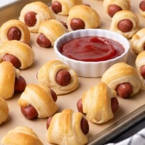 Completed pigs in a blanket are on a baking sheet with a ramekin of ketchup.