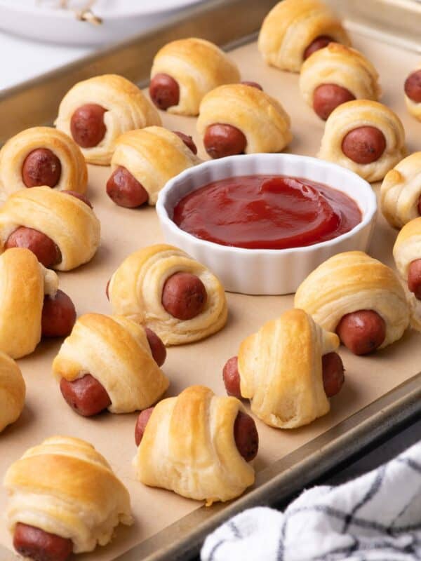 Completed pigs in a blanket are on a baking sheet with a ramekin of ketchup.