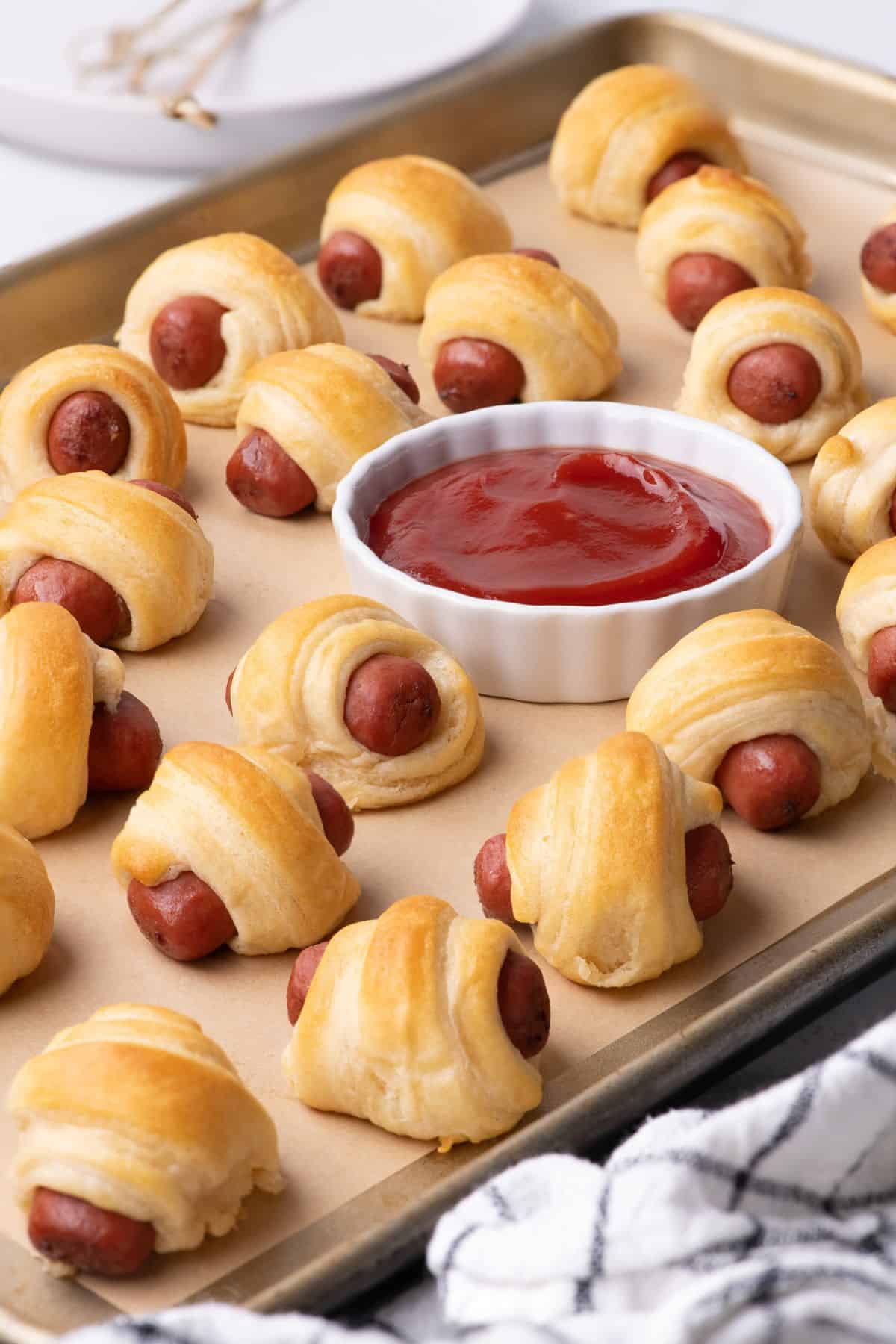 Completed pigs in a blanket are on a baking sheet with a ramekin of ketchup.