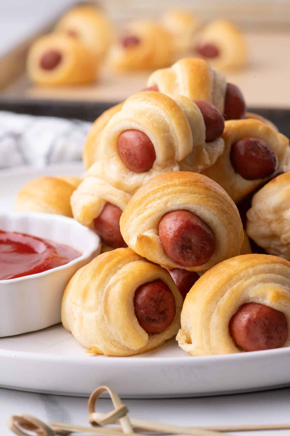 A large pile of pigs in a blanket are stacked next to a small bowl of ketchup on a white dinner plate.